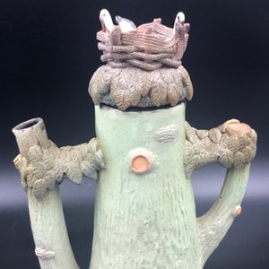 May include: A ceramic teapot shaped like a tree trunk with a bird's nest on top. The teapot has a green glaze and is decorated with leaves and branches. The bird's nest is made of twigs and has a white glaze.