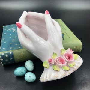 May include: White porcelain hands holding a small bowl, decorated with pink roses and green leaves. The hands have pink nail polish.