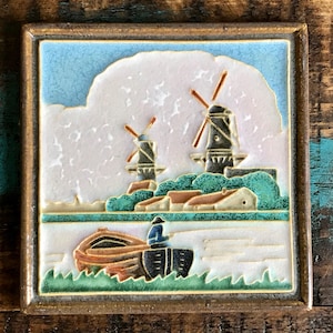 May include: Square ceramic tile with a Dutch landscape scene. The tile features two windmills, a boat with a person, and houses. The color palette includes blue, white, brown, green, and black, set within a gold-toned frame.