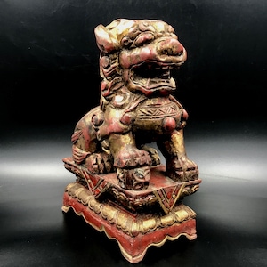 May include: A detailed, antique-style sculpture of a lion-like creature, predominantly red and gold. The figure is seated on a decorative base, showcasing intricate carvings and a weathered appearance. The artwork is a representation of traditional Asian art.