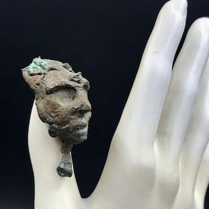 May include: A small, weathered bronze sculpture of a human face with a headdress. The sculpture has a detailed profile and is held against a white background. The bronze has a green patina.
