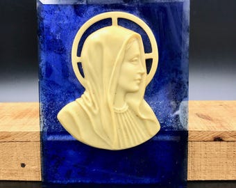 May include: A religious relief sculpture featuring a profile of a woman with a halo, set against a deep blue background. The woman's face and veil are a creamy off-white color. The artwork is mounted on a rectangular base.