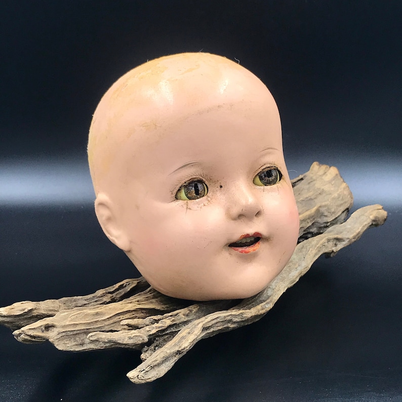 May include: A vintage porcelain doll head with painted green eyes and a slightly open mouth. The head is mounted on a piece of driftwood.