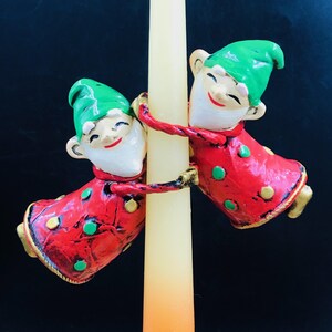 May include: Two ceramic elf figures with green hats and red coats featuring gold and green polka dots, each with a white beard and smiling expression, are attached to a tall, slender, pale yellow taper candle that stands vertically between them.