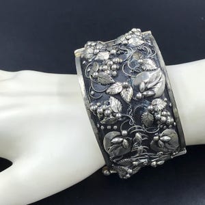 May include: A silver bracelet with an intricate design of leaves and grapes. The bracelet is wide and has a textured surface.