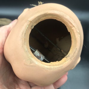 May include: Close-up of the back of a porcelain doll head, showing the opening for the doll's mechanism. The opening is circular and has a rough, unfinished edge. A metal rod is visible inside the opening.