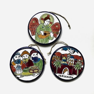 May include: Three round ceramic ornaments with a blue border and a white background. The first ornament depicts an angel playing a lute. The second ornament depicts three men carrying a tray of food. The third ornament depicts a cityscape with the word "Jerusalem" written below.