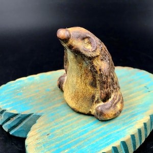 May include: A small, handcrafted ceramic badger figurine with a brown and tan color scheme. The badger is sitting upright, with its paws visible. The figurine is placed on a blue, wooden, leaf-shaped base.