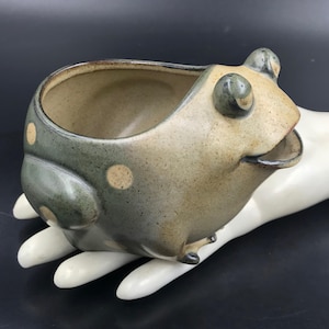 May include: A ceramic frog-shaped bowl with a speckled tan and muted green glaze. The vessel features a wide, open mouth, two prominent raised eyes on the top, and light-colored circular spots across the back and sides of the rounded body.