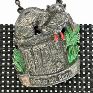 May include: A silver-colored metal ornament depicting a marten with red paws holding a bird in its mouth. The marten is sitting on a tree stump with green leaves. The ornament has the text "Marder mit Beute" on the bottom.