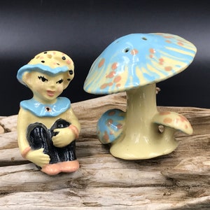 May include: A ceramic salt and pepper shaker set featuring a blue and yellow mushroom and a blue and yellow elf. The mushroom has a light blue top with brown spots and a yellow base. The elf is wearing a blue hat with white polka dots and a blue collar.
