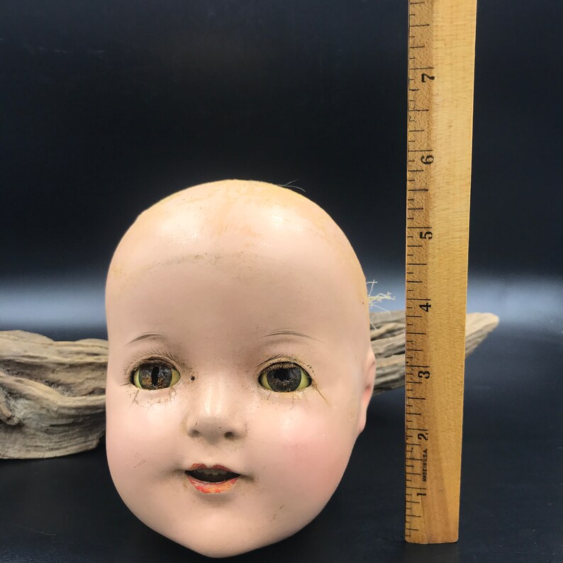 May include: A vintage porcelain doll head with painted brown eyes, a slightly open mouth, and a rosy complexion. The head is approximately 6 inches tall.