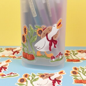 May include: A clear plastic pencil holder with a white goose sticker on the front. The goose is carrying a small brown basket and is surrounded by sunflowers. There are several other stickers of the same goose on a blue surface.