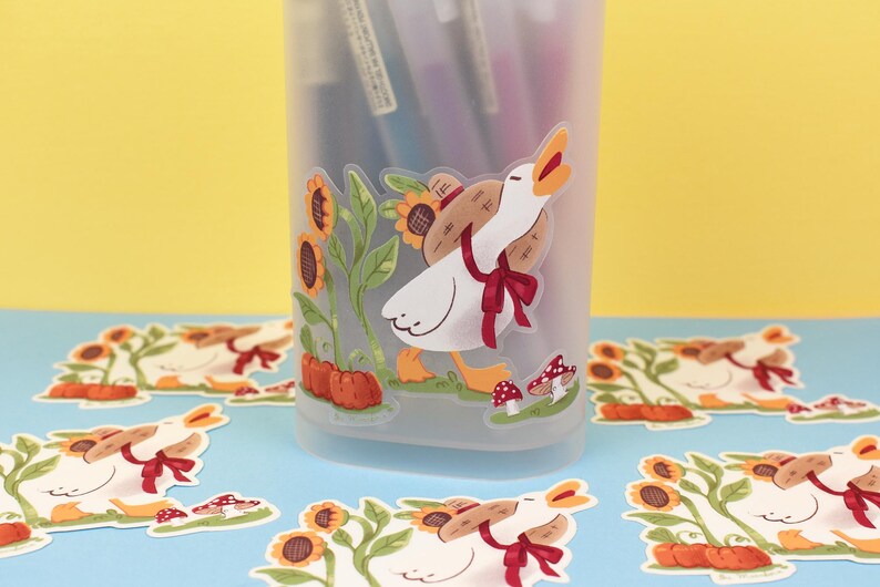 May include: A clear plastic pencil holder with a white goose sticker on the front. The goose is carrying a small brown basket and is surrounded by sunflowers. There are several other stickers of the same goose on a blue surface.