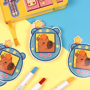 Pode incluir: Autocolantes com um personagem de urso de desenho animado dentro de um design de jogo port&aacute;til retro azul. O urso tem um corpo castanho, uma fruta laranja redonda na cabe&ccedil;a e um rosto sorridente. Um caderno amarelo e canetas coloridas tamb&eacute;m s&atilde;o vis&iacute;veis.