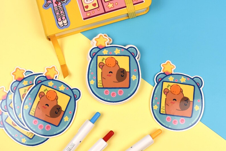 Pode incluir: Autocolantes com um personagem de urso de desenho animado dentro de um design de jogo port&aacute;til retro azul. O urso tem um corpo castanho, uma fruta laranja redonda na cabe&ccedil;a e um rosto sorridente. Um caderno amarelo e canetas coloridas tamb&eacute;m s&atilde;o vis&iacute;veis.