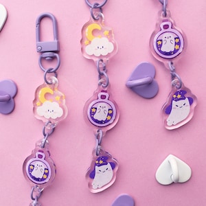 Puede incluir: Un conjunto de cinco llaveros con dijes de color morado y blanco con diferentes diseños. Los dijes incluyen un fantasma en una botella, un fantasma con un sombrero de bruja y una nube con una luna y estrellas. Los llaveros están unidos a anillos en forma de corazón morado.