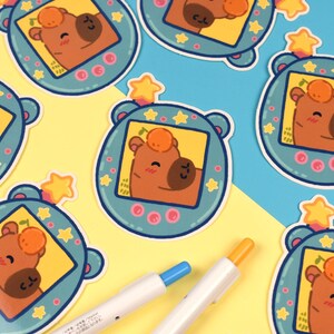 Pode incluir: Uma cole&ccedil;&atilde;o de autocolantes com um personagem de urso de desenho animado dentro de um dispositivo azul-petr&oacute;leo com uma estrela no topo. O urso &eacute; castanho com uma pequena fruta laranja na cabe&ccedil;a. Duas canetas com pontas azuis e laranja est&atilde;o em primeiro plano.