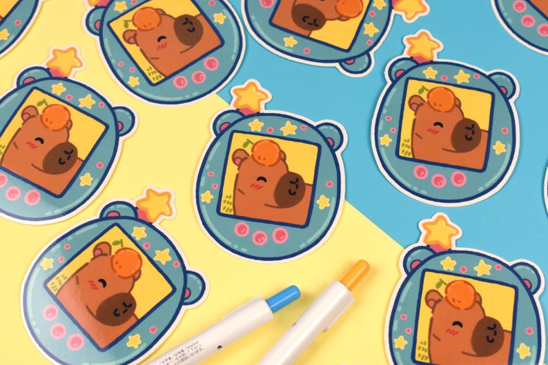 Pode incluir: Uma cole&ccedil;&atilde;o de autocolantes com um personagem de urso de desenho animado dentro de um dispositivo azul-petr&oacute;leo com uma estrela no topo. O urso &eacute; castanho com uma pequena fruta laranja na cabe&ccedil;a. Duas canetas com pontas azuis e laranja est&atilde;o em primeiro plano.