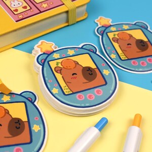 Pode incluir: Adesivos azuis com um desenho de urso de desenho animado, apresentando um urso marrom com uma fruta laranja na cabe&ccedil;a. Os adesivos est&atilde;o em um fundo amarelo e azul, com um caderno amarelo e canetas.