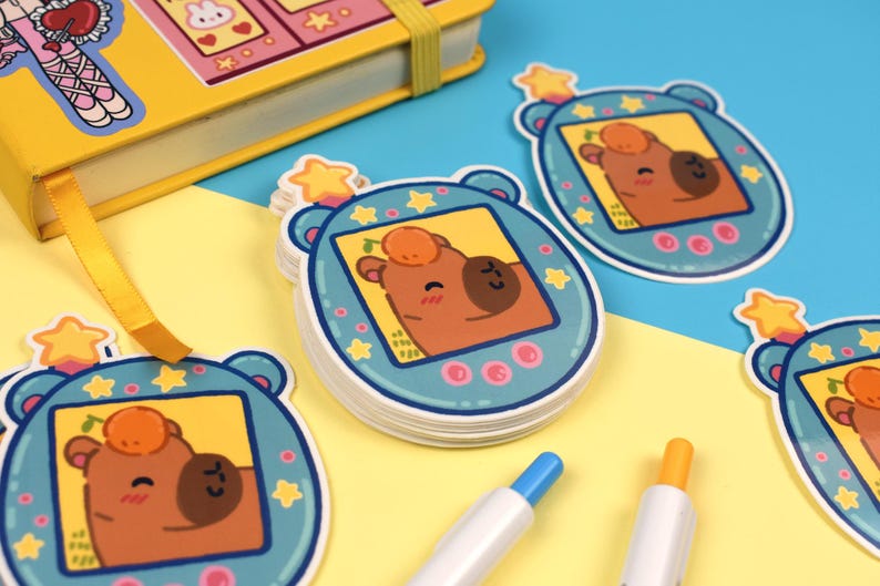 Pode incluir: Adesivos azuis com um desenho de urso de desenho animado, apresentando um urso marrom com uma fruta laranja na cabe&ccedil;a. Os adesivos est&atilde;o em um fundo amarelo e azul, com um caderno amarelo e canetas.
