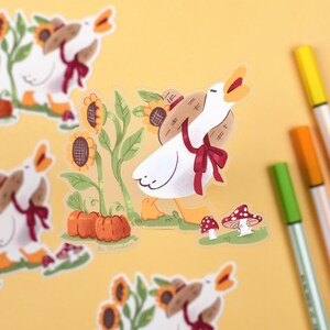 May include: A set of five stickers featuring a white goose carrying a basket of pumpkins and sunflowers. The stickers are on a yellow background with markers.