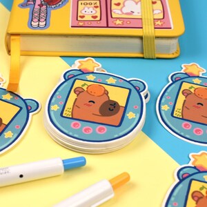 Pode incluir: Uma pilha de adesivos com um personagem de urso de desenho animado em uma moldura azul com uma estrela no topo. Os adesivos est&atilde;o em uma superf&iacute;cie amarela e azul, com um caderno amarelo e canetas no fundo. O urso tem um rosto marrom e uma pequena fruta laranja.