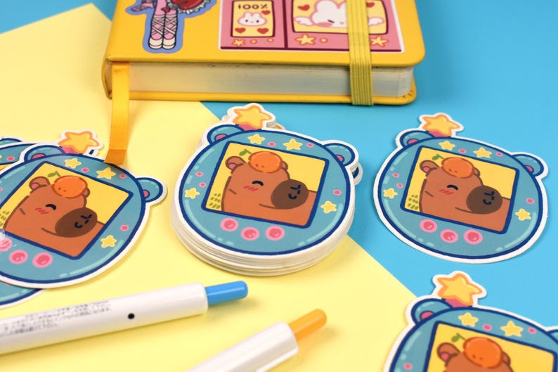 Pode incluir: Uma pilha de adesivos com um personagem de urso de desenho animado em uma moldura azul com uma estrela no topo. Os adesivos est&atilde;o em uma superf&iacute;cie amarela e azul, com um caderno amarelo e canetas no fundo. O urso tem um rosto marrom e uma pequena fruta laranja.
