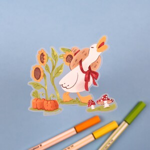 May include: A white goose with a red bow and a straw hat is walking towards a patch of sunflowers and pumpkins. The goose is carrying a basket of sunflowers on its back.