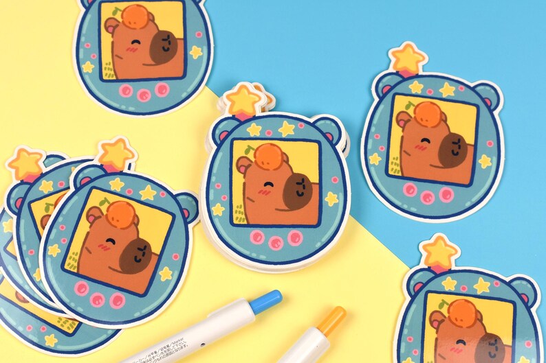 Pode incluir: Uma cole&ccedil;&atilde;o de adesivos com um personagem de urso de desenho animado dentro de um dispositivo de jogo port&aacute;til azul em estilo retr&ocirc;. O urso tem um rosto marrom e uma fruta laranja na cabe&ccedil;a. Os adesivos est&atilde;o em um fundo amarelo e azul.