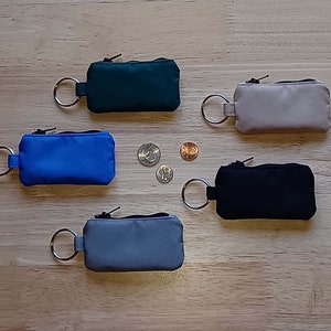 May include: Five rectangular zippered pouches with attached metal key rings in dark green, beige, blue, black, and gray. The pouches are arranged around three small coins placed in the center, showing their compact size and functional design.