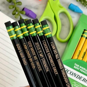 Puede incluir: Un conjunto de lápices negros con letras doradas, cada uno con un mensaje inspirador. Los lápices tienen detalles verdes y amarillos. También hay tijeras verdes, gomas de borrar y una caja de lápices.