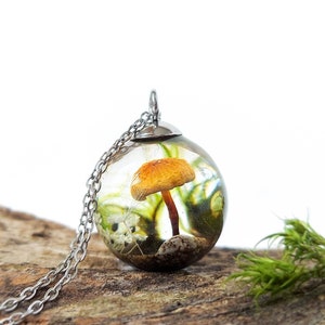 On a wooden log with a white background, a crystal resin sphere pendant. Lovely soft brown mushroom inside surrounded by real green moss, a dandelion seed, a small white flower and a shell, all on a sand base. Stainless steel cap and chain necklace.