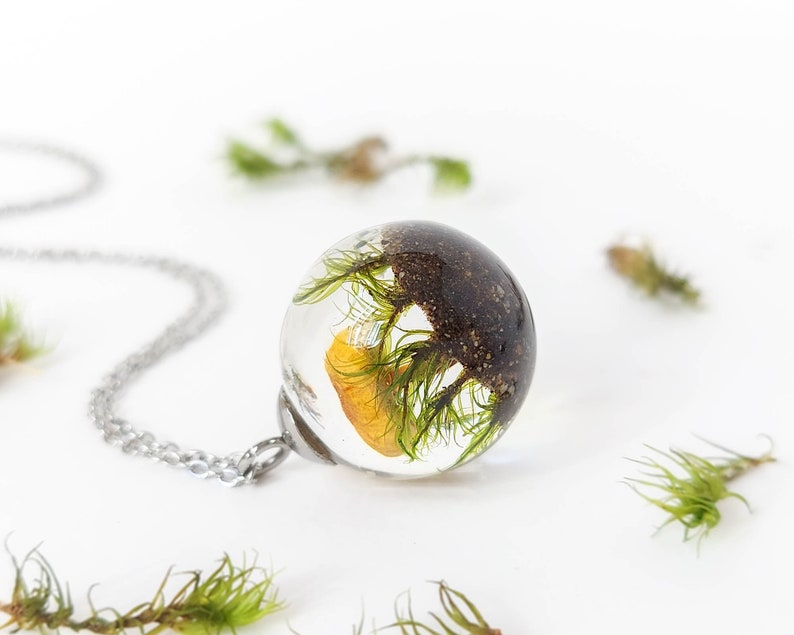 Beautiful spherical glass pendant made of transparent resin. View of its rear part where you can see in detail the natural green moss on sand with the mushroom in the background. Shiny chain made of hypoallergenic stainless steel.