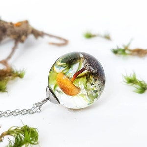 Cottagecore style pendant handmade with true elements of nature: A mushroom, a dandelion seed, a conch shell, a small white flower; all surrounded by natural moss on sand. The cap and the chain are made of stainless steel. Luminous and crystalline!