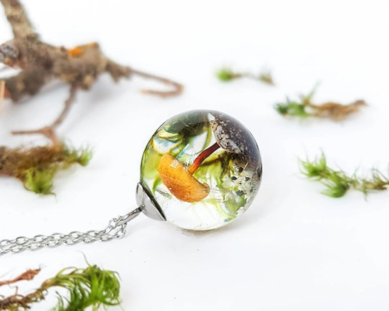 Cottagecore style pendant handmade with true elements of nature: A mushroom, a dandelion seed, a conch shell, a small white flower; all surrounded by natural moss on sand. The cap and the chain are made of stainless steel. Luminous and crystalline!