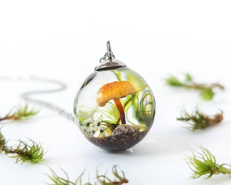 Necklace with a crystal resin sphere pendant on a white background and real mosses. Magical forest inspiration in whose interior a real light brown mushroom stands out with a dandelion seed, a small white flower, and a shell among real moss.