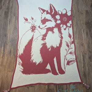 Könnte beinhalten: Eine rechteckige cremefarbene Decke mit einem roten Fuchsmotiv und floralen Akzenten. Der Fuchs sitzt, mit einer Blume hinter seinem Kopf. Die Decke hat einen roten Rand und Quasten an jeder Ecke. Der Text "The Enchanted Weaver" befindet sich unten.