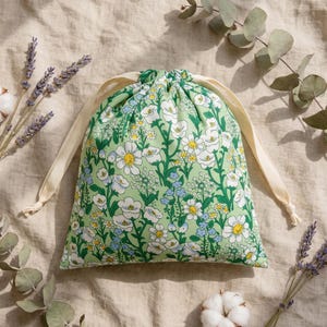 May include: A light green drawstring pouch featuring a repeating pattern of white, yellow, and blue wildflowers. The bag is constructed with cream-coloured fabric ribbons that cinch the top closed, creating a gathered opening for storage.