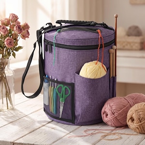 May include: A cylindrical purple yarn storage bag with a black handle and shoulder strap. The bag features exterior mesh pockets containing crochet hooks and scissors, a side pocket holding a ball of yellow yarn, and slots for knitting needles.