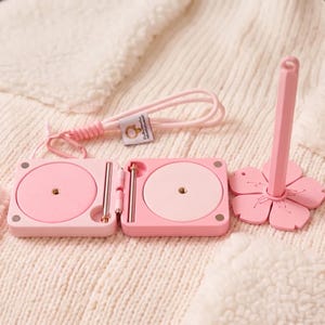 Könnte beinhalten: Ein hellrosa Klappetui mit zwei runden Fächern, die jeweils einen Metallstift enthalten. Eine passende rosa Kordel mit einem kleinen Etikett ist befestigt. Neben dem Etui steht ein blumenförmiger Ständer mit einem vertikalen Stab.