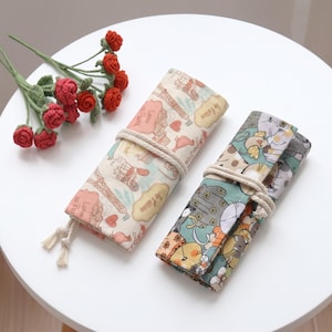 May include: Two fabric roll-up pencil cases secured with white rope ties. One case features a cream background with Paris-themed illustrations including the Eiffel Tower and hearts. The other case displays a colorful pattern of cartoon cats.