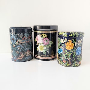 May include: Three black metal canisters with floral designs. One features a pattern of birds and foliage, another has a bouquet with the text "PEONY ROSE ANEMONE", and the third displays colorful flowers. All have black lids.