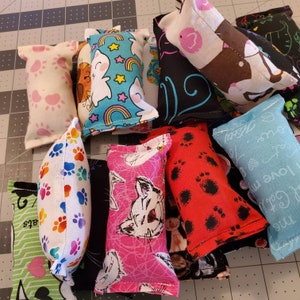 May include: A collection of cat toys in various colors and patterns. The toys are rectangular and made of fabric, with designs including paw prints, cats, rainbows, and swirls. Some toys are pink, red, blue, and white.