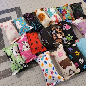 May include: A collection of cat toys in various colors and patterns. Designs include paw prints, cats, and whimsical graphics. The toys are rectangular and filled, perfect for feline entertainment.
