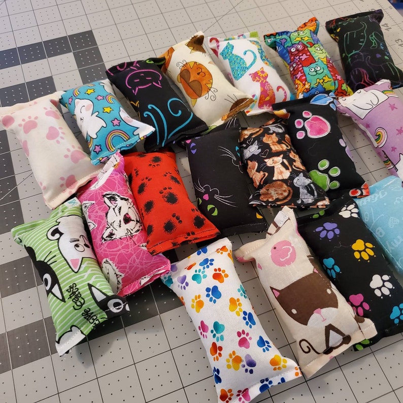 May include: A collection of cat toys in various colors and patterns. Designs include paw prints, cats, and whimsical graphics. The toys are rectangular and filled, perfect for feline entertainment.