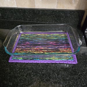 May include: A clear glass baking dish sits on a purple and black patterned hot pad. The hot pad has a black background with colorful streaks of rainbow colors. The edges of the hot pad are purple with small white dots.