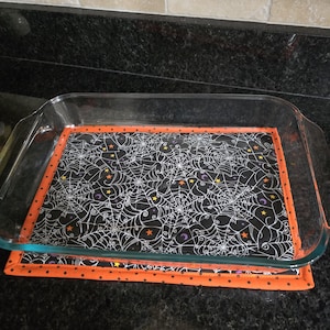 May include: A clear glass baking dish rests on a Halloween-themed hot pad. The hot pad features a black background with white spiderwebs, orange trim, and orange polka dots. The dish is on a black countertop.