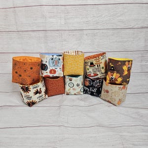 May include: A collection of 10 small fabric storage bins with various autumnal designs. The bins feature pumpkins, turkeys, fall leaves, and the words "Happy Fall Y'all" and "Gobble 'til you wobble".