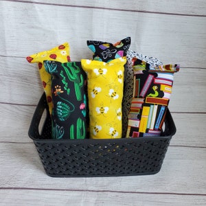 May include: A black wicker basket filled with five colorful fabric-covered pillows. The pillows have different patterns, including cacti, bees, and books.